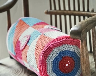 May include: A colorful, handmade crochet pillow in a cylindrical shape. The pillow features a patchwork design with shades of pink, orange, blue, and white. It rests on a wooden chair, showcasing its vibrant, textured surface.