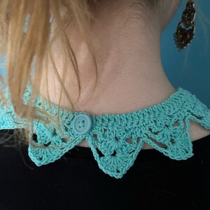 May include: A turquoise crocheted collar with a button closure. The collar has a scalloped edge and is made with a delicate stitch.
