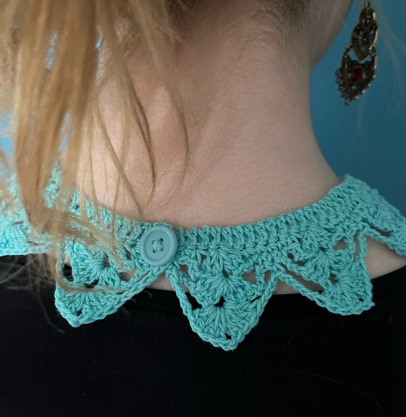 May include: A turquoise crocheted collar with a button closure. The collar has a scalloped edge and is made with a delicate stitch.