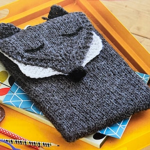 May include: A gray knitted phone case shaped like a wolf's head with a white felt nose and white felt ears. The case is lying on a yellow surface with a blue and white patterned book underneath.