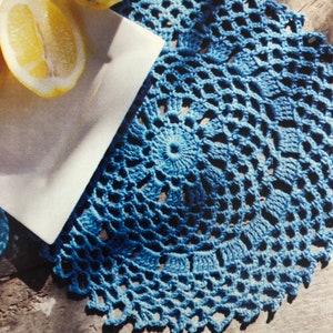 Könnte beinhalten: Eine blaue gehäkelte Deckchen mit einem runden Design. Die Deckchen hat ein zartes, spitzenartiges Muster und ist perfekt, um jedem Tisch eine elegante Note zu verleihen.