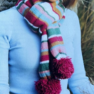 以下が含まれることがあります： ピンク、白、茶色、緑のストライプが入ったストライプのストール。ストールの両端には大きなポンポンが2つ付いており、どちらも濃いワインレッドの色です。