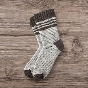 May include: A pair of crocheted socks in light grey with dark grey details on the toes, heels, and cuffs. The cuffs have horizontal stripes. The socks are presented on a wooden surface.