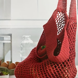 Pode incluir: Uma sacola de compras vermelha de crochê com alças longas, cheia de produtos. A sacola tem um design de rede e está em cima de uma mesa. Um pimentão vermelho é visível dentro da sacola, junto com outros produtos.