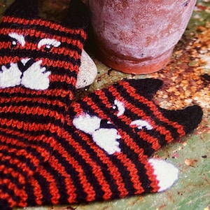 Könnte beinhalten: Zwei gestrickte Katzenhandschuhe in Schwarz und Rot mit weißen Filzgesichtern. Die Handschuhe liegen auf einer grün-braunen Oberfläche.
