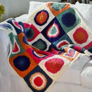 Puede incluir: Una colorida manta de ganchillo hecha a mano con un patrón geométrico. La manta presenta cuadrados con círculos en tonos naranja, rosa, azul, verde y blanco. La manta está sobre una superficie blanca.