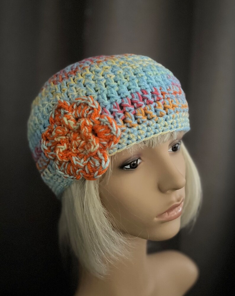 May include: A crocheted hat with a flower design in shades of blue, orange, and yellow. The hat is made with a soft, textured yarn and has a slouchy fit.
