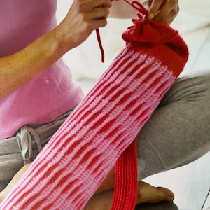 Może przedstawiać: Różowo-czerwony, dziergany na szydełku pokrowiec na matę do jogi ze sznurkiem do ściągania. Pokrowiec trzyma osoba ubrana w różową koszulę i szare spodnie.