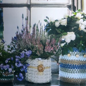 May include: Three potted plants with crocheted covers. The plant on the left has blue flowers, the middle plant has pink flowers, and the plant on the right has white flowers. The crocheted covers are in shades of blue, white, and brown.