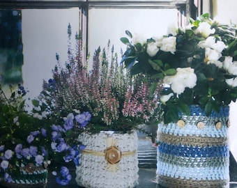May include: Three potted plants with crocheted covers. The plant on the left has blue flowers, the middle plant has pink flowers, and the plant on the right has white flowers. The crocheted covers are in shades of blue, white, and brown.