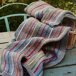 May include: A gray striped knitted throw blanket with colorful stripes in red, yellow, blue, green, and white. The blanket is folded and draped over a wooden surface.
