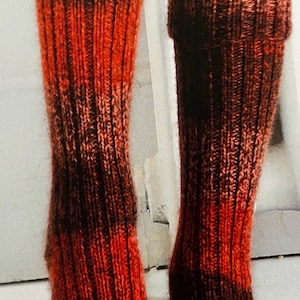 Puede incluir: Un par de calcetines largos de punto con un patrón degradado rojo y marrón. Los calcetines están hechos con un hilo grueso y tienen una textura acanalada.