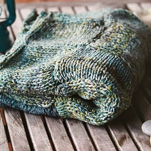 May include: A folded knitted blanket in shades of blue, green, and white. The blanket is made with a textured stitch and has a soft, cozy feel.