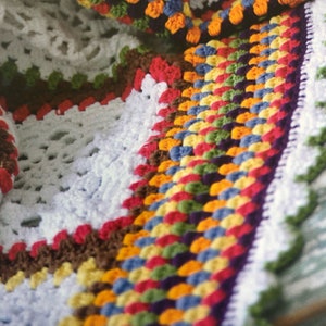May include: A close-up of a colorful crocheted blanket with a white background. The blanket is made with a variety of colors, including red, orange, yellow, green, blue, and purple. The blanket is crocheted in a granny square pattern.