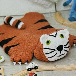 May include: A plush tiger rug with a white face, black stripes, and green eyes. The rug is lying on a wooden floor with toy cars and a train track.