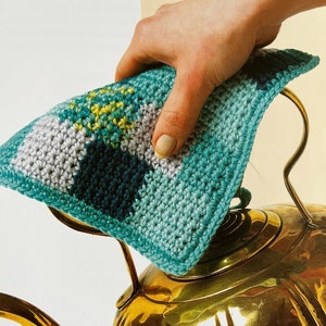 May include: A crocheted pot holder in shades of teal, green, and gray with a geometric pattern. The pot holder is being used to protect a gold teapot.