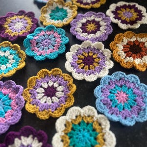 Puede incluir: Una colección de 12 posavasos de ganchillo en varios colores y patrones, incluyendo morado, azul, rosa, amarillo y blanco. Los posavasos son todos circulares y tienen un diseño floral.