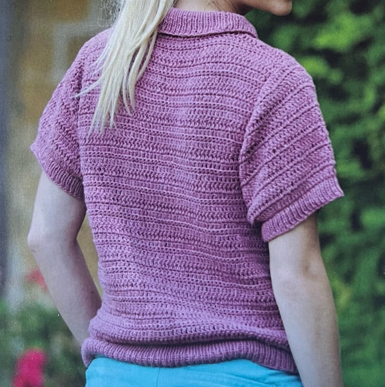 Puede incluir: Una camisa de punto de crochet de color rosa con mangas cortas y cuello.