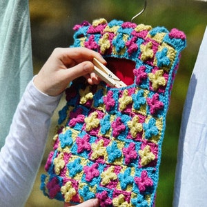 May include: A crocheted hanging organizer with a floral pattern in shades of pink, yellow, and blue. The organizer has a pocket for storing items and is hanging from a wooden clothes peg.