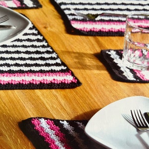 Könnte beinhalten: Vier gehäkelte Tischsets mit schwarzen und weißen Streifen und rosa Einfassung. Die Tischsets liegen auf einem Holztisch mit einem weißen Teller und einem Glas Wasser.