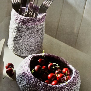 May include: A crocheted utensil holder and bowl. The utensil holder is cylindrical and holds several forks. The bowl is filled with ripe red cherries. Both items are made of a gray and purple speckled yarn.