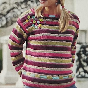 May include: A striped jumper with a colourful button embellishment on the shoulder. The jumper is made in a variety of colours, including pink, green, purple, and white.