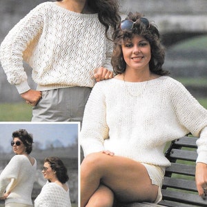 May include: Two women wearing white knitted sweaters with openwork patterns. One woman is standing with her hands in her pockets, while the other is sitting on a wooden bench.