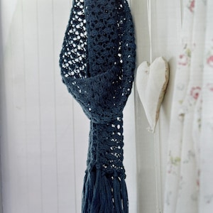May include: A dark blue, open-weave crochet scarf with a fringed bottom hangs from a white hook. The scarf has a textured appearance and a soft drape. A wooden heart and floral curtain are in the background.
