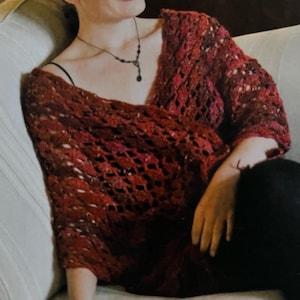 May include: A burgundy crochet sweater with a loose, open weave. The sweater has long sleeves and a deep V-neck. The garment is worn over a black camisole and paired with black pants. A delicate necklace with a pendant is also visible.