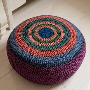 May include: A circular crocheted pouffe with a colourful design. The pouffe features concentric rings of colour, including blue, orange, green, and purple. A decorative and functional item for the home.
