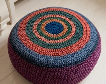 May include: A round, crocheted pouf in a vibrant color palette. The design features concentric circles of color, including blue, orange, green, and purple. The pouf is a decorative and functional piece of home decor.