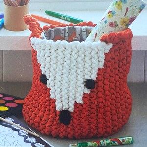 May include: A knitted fox-shaped basket with a white face and orange body. The basket is filled with colorful patterned paper.