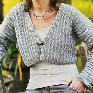 May include: A gray cropped cardigan with a V-neck and a wooden button closure. The cardigan has a ribbed texture and is worn over a white tank top.