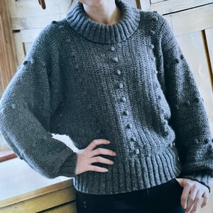 May include: A grey knitted jumper with a round neckline and a textured pattern. The jumper has a loose fit and long sleeves.
