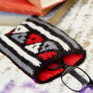 May include: A black, white, and red knitted glasses case with a geometric pattern. The case is open and a pair of glasses is inside.