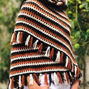 May include: A handmade crochet poncho in black, white, and brown stripes with fringe. The poncho features a cowl neck and is made with a textured stitch. The fringe is a mix of brown and white tassels, adding a bohemian flair to the garment.