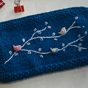 May include: A rectangular, dark blue knitted pouch featuring white embroidered branches and three small, colorful bird-shaped buttons in pink, light green, and blue. The pouch has a white zipper closure along the top edge and a textured knit finish.