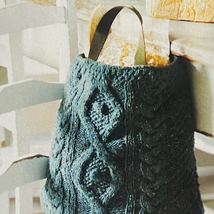 Puede incluir: Una bolsa de mano de punto azul verdoso con un asa de cuero marrón. La bolsa está llena de pan y está sentada en una silla blanca.