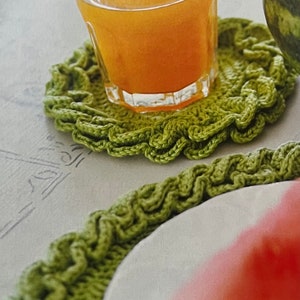May include: A glass of orange juice sits on a green crocheted coaster. The coaster has a ruffled edge.