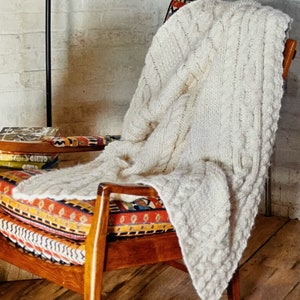 Puede incluir: Una manta de punto de cable blanco drapeada sobre un sillón de madera con un cojín de estampado colorido. La silla está en una habitación con una pared de ladrillo blanco y un suelo de madera.