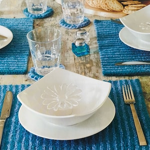 May include: A white ceramic bowl with a decorative design sits on a white plate. The bowl and plate are on a blue and white striped placemat. A silver fork is on the right side of the image.