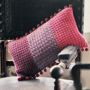 May include: A rectangular, two-toned crochet cushion in shades of pink and purple, trimmed with red pom-poms. The cushion is placed on a vintage chair, highlighting its textured surface and decorative design.