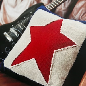 May include: A white and blue knitted pillow with a large red star in the center. The star is outlined in white and has a slightly textured surface.