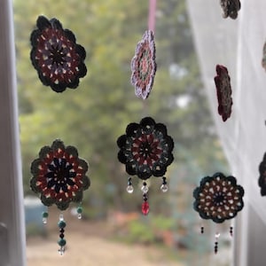 May include: Crochet flower decorations in shades of black, red, and green, some with dangling beads. The handmade ornaments are suspended in front of a window, creating a decorative display. The crochet flowers are a unique craft.
