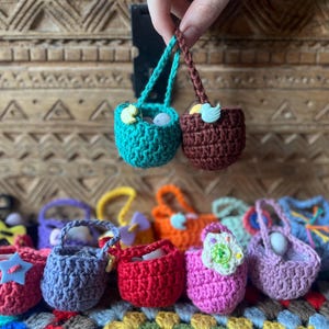 Può includere: Una collezione di 10 piccoli cestini all'uncinetto in vari colori, tra cui viola, rosa, rosso, verde e marrone. I cestini sono decorati con bottoni e altri abbellimenti.