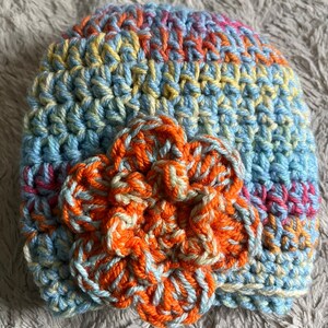 May include: A crocheted hat with a flower design. The hat is made with blue, yellow, orange, and pink yarn. The flower is made with orange and white yarn.