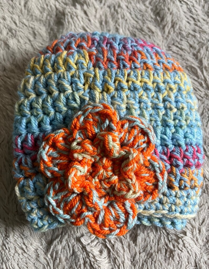 May include: A crocheted hat with a flower design. The hat is made with blue, yellow, orange, and pink yarn. The flower is made with orange and white yarn.