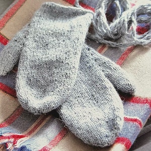 May include: A pair of gray knitted mittens with a textured pattern. The mittens are lying on a plaid fabric with red, blue, and white stripes.