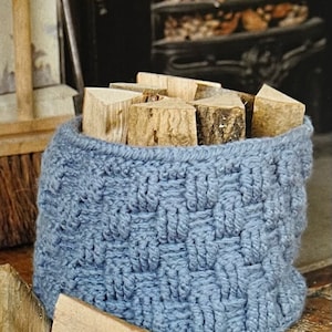 May include: A blue crocheted basket filled with stacked pieces of firewood. The basket is sitting on a wooden surface.