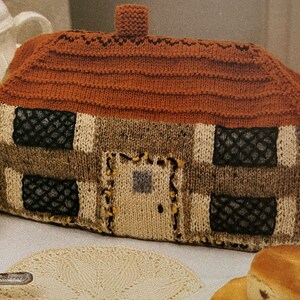 Op de afbeelding: Een gebreide theepotwarmer in de vorm van een huis met een bruin dak, witte en bruine muren en zwarte ramen.